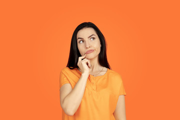 Caucasian young woman's portrait on orange studio background. Beautiful female brunette model in shirt. Concept of human emotions, facial expression, sales, ad. Copyspace. Dreamful, thoughtful.