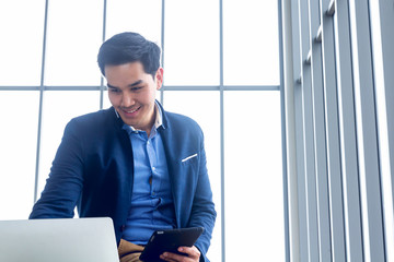 Young businessman wearing a suit and a long-sleeve shirt inside and sitting smart poses in a modern office. In his hand have a tablet in action looking laptop screen for check email reading the news 