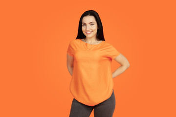 Obraz premium Caucasian young woman's portrait on orange studio background. Beautiful female brunette model in shirt. Concept of human emotions, facial expression, sales, ad. Copyspace. Smiling, looks confident.