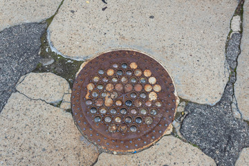 Rusted iron and glass manhole cover set in crumbling concrete and asphalt sidewalk, horizontal aspect