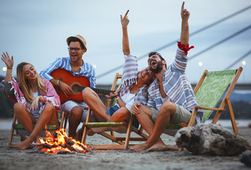 Group of friends sitting around camp fire at the beach at the autumn evening.They play guitar and singing.