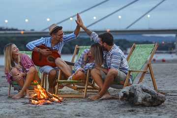 Group of friends sitting around camp fire at the beach at the autumn evening.They play guitar and singing.