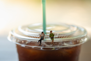 Business and communication concept. businessman with newspaper and woman miniature figures sitting and talking on top of cover lid of plastic cup of iced black coffee (Americano)