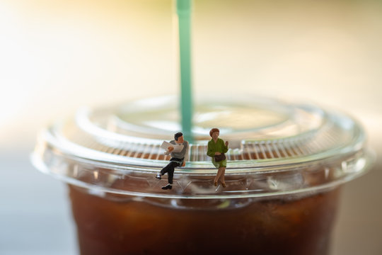 Business And Communication Concept. Businessman With Newspaper And Woman Miniature Figures Sitting And Talking On Top Of Cover Lid Of Plastic Cup Of Iced Black Coffee (Americano)