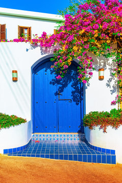 Traditional Mediterranean House With A Stone Fence And A Large Blue Door Decorated With Flowering Plants. Front View