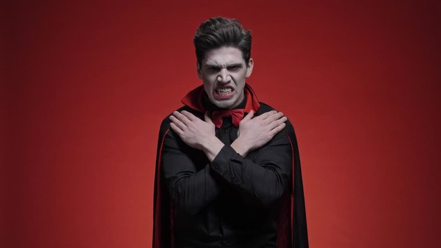 Terrible man dracula with crossed hands in black halloween costume is roaring isolated over red wall                                                                                  