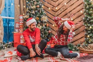 Red-dressed people are playing around. Girl is moking up, holding candycanes above her head, imitating a reindeer. Her man is laughing desparately.