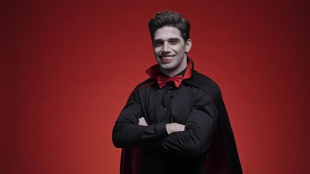 Vampire man with blood and fangs in black halloween costume is winking and smiling while looking at the camera over red wall isolated