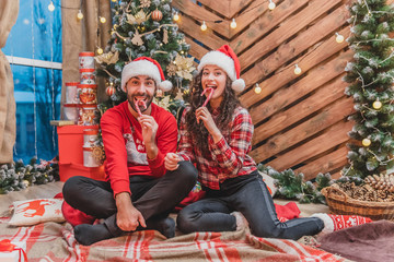 Full-body photo of young people in red clothes and Santa hats, tasting yummy candycanes in christmas atmosphere.