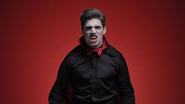 Angry vampire man with blood and fangs in black halloween costume screaming  and looking at the camera isolated over red wall