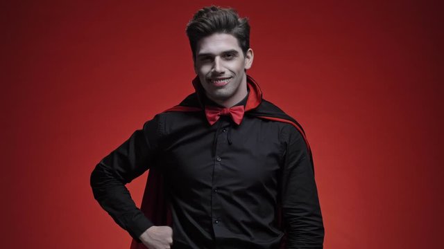 Happy vampire man with blood and fangs in black halloween costume gesturing at camera isolated over red wall