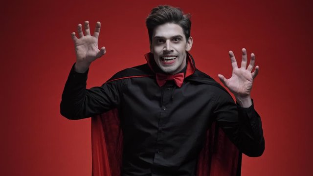 Vampire man with blood and fangs in black halloween costume appearing with scary gesture over red wall