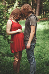 Fototapeta premium A happy married couple Husband hugs a beautiful adult pregnant wife. walks in a park. Second trimester. Holds his hands on her stomach. The benefits of walking during pregnancy, health care.