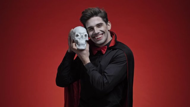 Glad vampire man with fangs in black halloween costume holding human skull and stroking it isolated over red wall
