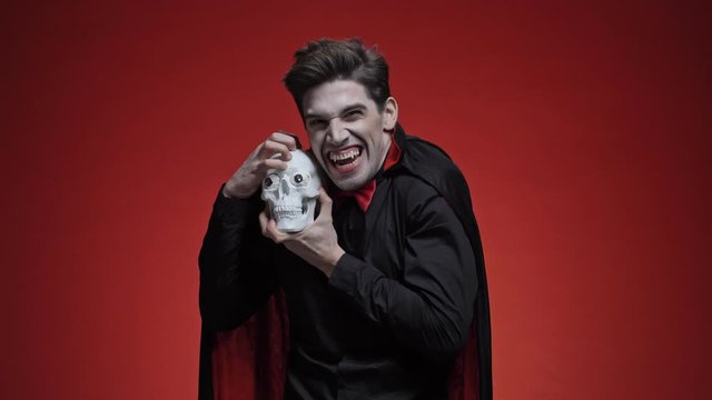 Scary vampire man with fangs in black halloween costume looking at human skull and roaring with it to the camera isolated over red wall