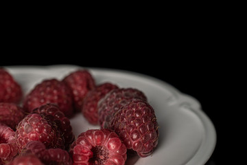 Lot of whole fresh crimson raspberry on white plate isolated on black glass