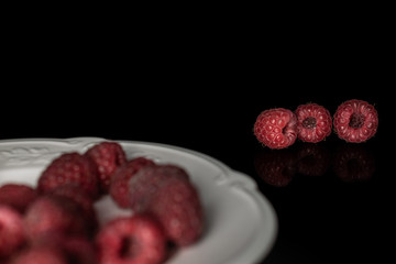Lot of whole fresh crimson raspberry on white plate isolated on black glass