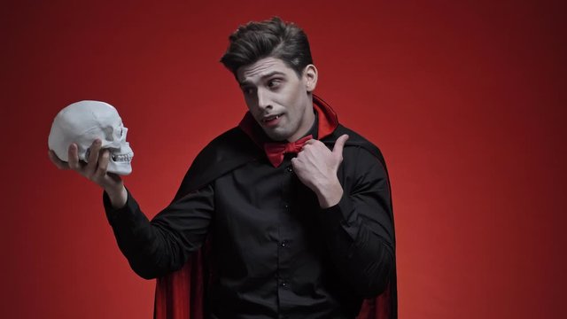 Scary vampire man with fangs in black halloween costume holding human skull in hand and talk to it isolated over red wall