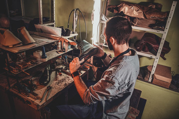 Diligent cobbler in glasses is working with pair of leather shoes at his workplace.