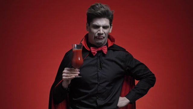 Scary vampire man with fangs in black halloween costume tasting a tomato cocktail and disliking it isolated over red wall