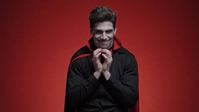 Mystic vampire man with blood and fangs in black halloween costume is planning something isolated over red wall