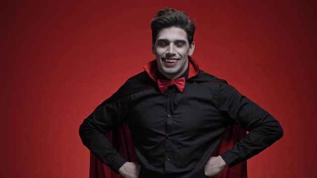 Handsome vampire man with fangs in black halloween costume shaking his head approvingly isolated over red wall