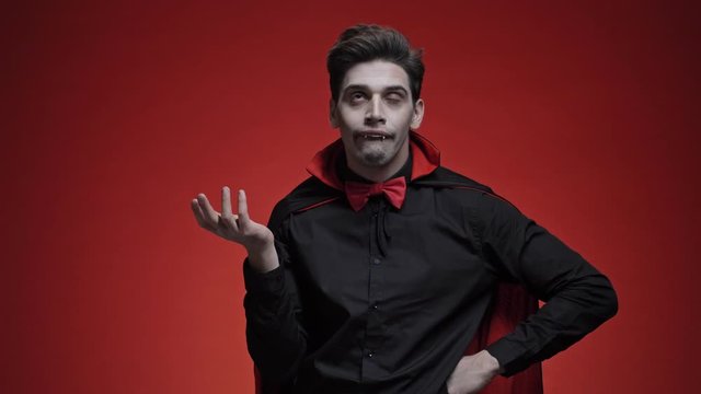 Vampire man with fangs in black Halloween costume rolling his eyes and feeling bored isolated over red wall