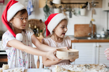 Children are cooking Christmas cookies in cozy home kitchen. Cute kids prepare holiday food for family. Funny little sisters in flour make New Year meal. Children chef concept. Lifestyle candid moment