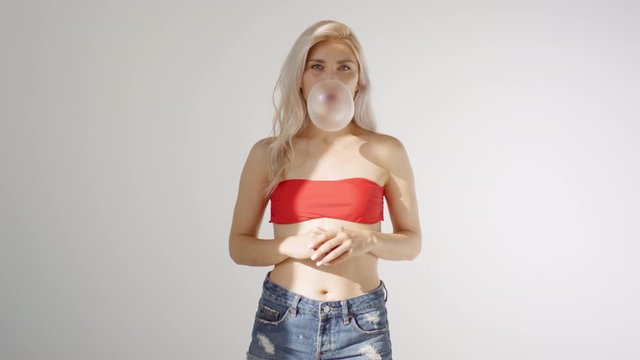 Thigh-up shot of young Caucasian woman with long blonde hair, in strapless crop top and denim shorts, standing against white wall, blowing bubble from chewing gum, then bubble bursting onto her face