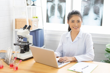 Asian female doctor working on laptop with a microscope for an exam at hospital giving patient convenience online service advice, smiling write a prescription , healthcare, preventing disease concept