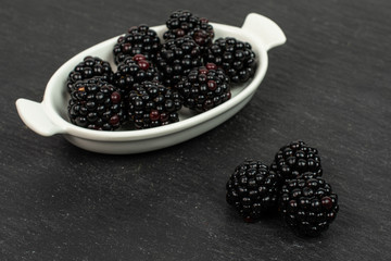 Lot of whole fresh black blackberry in white oval ceramic bowl on grey stone