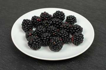 Lot of whole fresh black blackberry on white ceramic plate on grey stone