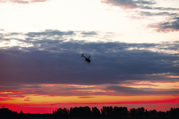 Silhouette of a chopper high in the evening sky at sundown seen from below, the line of the forest in contre-jour.