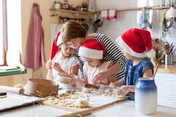 Family is cooking Christmas gingerbread cookies in cozy home kitchen for holiday dinner. Funny kids and woman bake Xmas biscuits. Mother is teaching daughters. Lifestyle moment. Children chef concept.