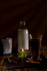Fototapeta na wymiar Glass of coffee on a wooden table with a bottle of milk, sugar bags, cologne and spices lit by candles.