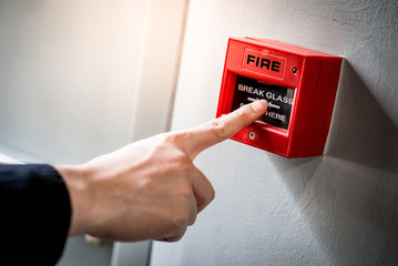 Male hand pointing at red fire alarm switch on concrete wall in office building. Industrial fire...
