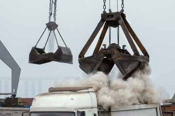 Grapple crane unloading cement clinker from ship to truck in river port. Barge delivery of dusty...
