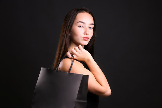 Black friday sale concept. Attractive young woman with long brunette hair, smiling, wearing sexy dress, holding a single blank shopping bag over black paper textured background. Copy space, close up.