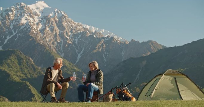 Old caucasian couple taking a rest on top of mountain, sitting and rehydrating while enjoying the view, talking. Mature people spending time together after retirement travelling - tourism, pension