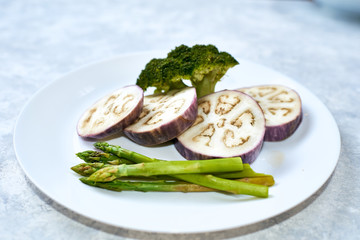 Healthy eating concept. Steamed vegetables in a white plate on a blue table. Eggplant, broccoli, asparagus.