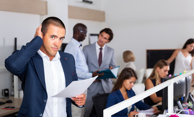 Puzzled businessman standing with papers in hands in open plan office