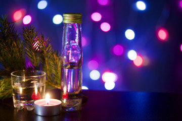 Obraz na płótnie Canvas Burning candle with a drink on the background with colorful lights. Nearby is a wineglass, a bottle of drink and a fir branch. Holiday accessories at dusk.