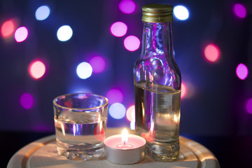 Burning candle with a drink on the background with colorful lights. Nearby is a glass and a bottle of drink. Holiday accessories at dusk.