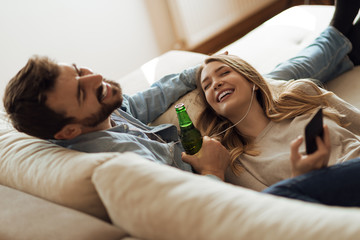 Young happy couple drinking beer and listening music on earphones at home