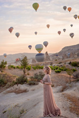 woman is watching on scenery view with rising balloons on sunrise. Girl in gorgeous pink long dress dance on hill looking at large number of air balls. Fabulous Cappadocia mountains landscapes Turkey