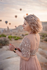 woman is watching on scenery view with rising balloons on sunrise. Girl in gorgeous pink long dress stay on hill looking at large number of air balls. Fabulous Cappadocia mountains landscapes Turkey