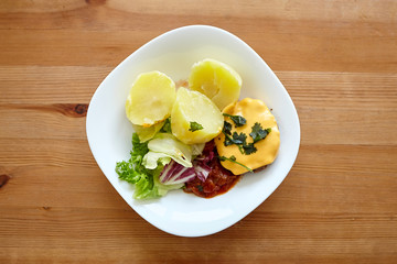 Boiled potatoes with green salad, vegetables and cutlet. White plate with homemade food on a wooden dinner table. Cooked potato dish on wooden background, top view