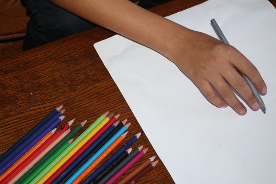 Hands of the artist close-up, holds a flamaster over a blank sheet of paper near scattered colored felt-tippers on a brown wooden table. View from above. Copy space