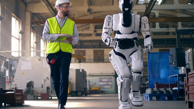 Man watches a robot walk in a factory room.