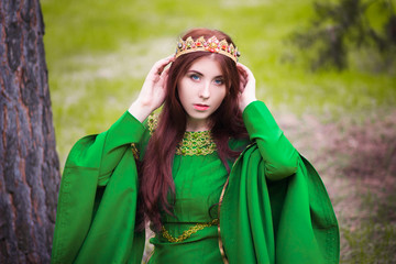 Beautiful, young, red-haired girl in a green medieval dress with long sleeves, with a golden crown, sits on the grass, in a fairy forest. Fantastic photosession. A model with clean skin.